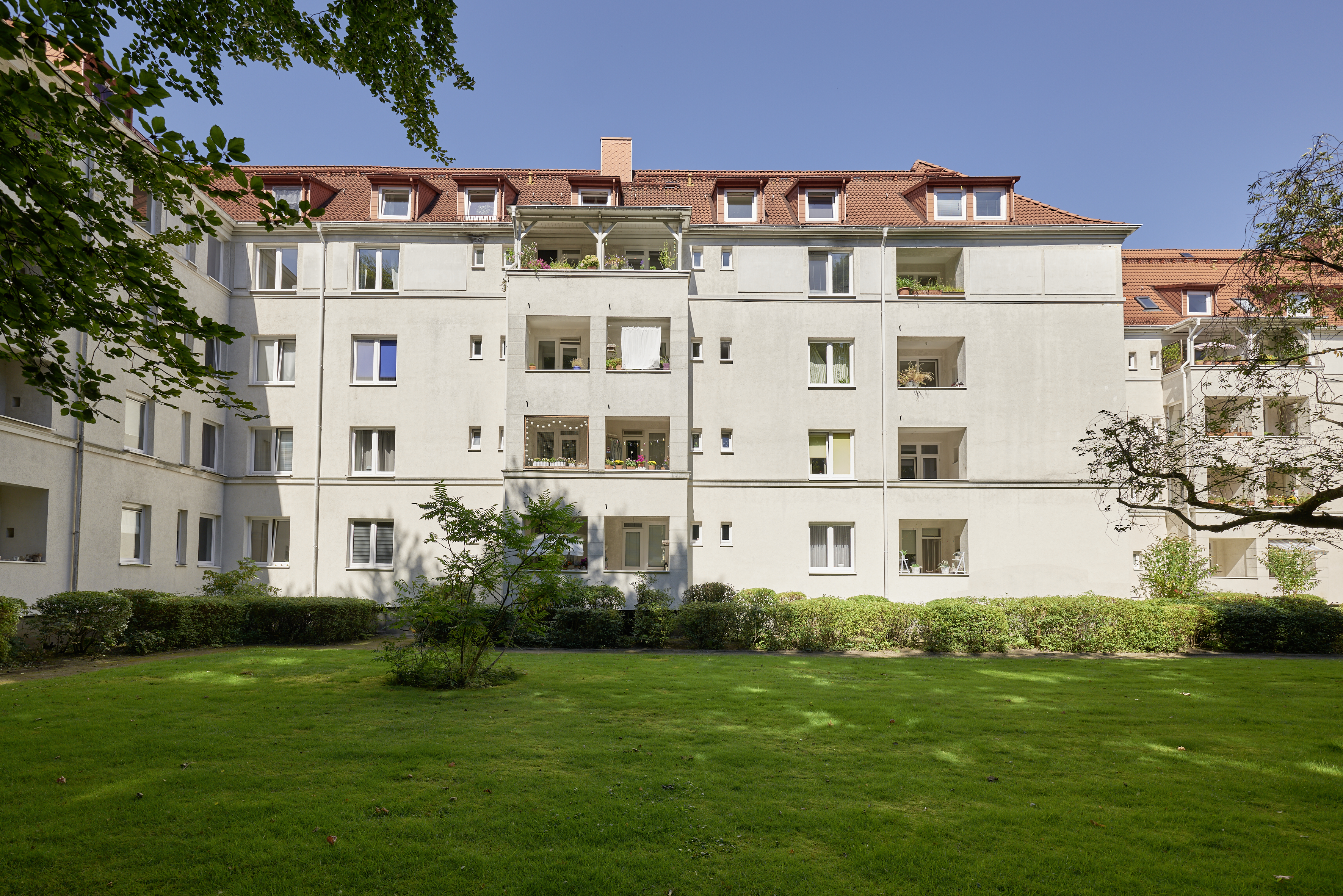 Viergeschossiges Wohnhaus mit heller Fassade, rotem Ziegeldach und Balkonen. Im Vordergrund liegt eine begrünte Innenhoffläche mit Rasen, Hecken und Bäumen.