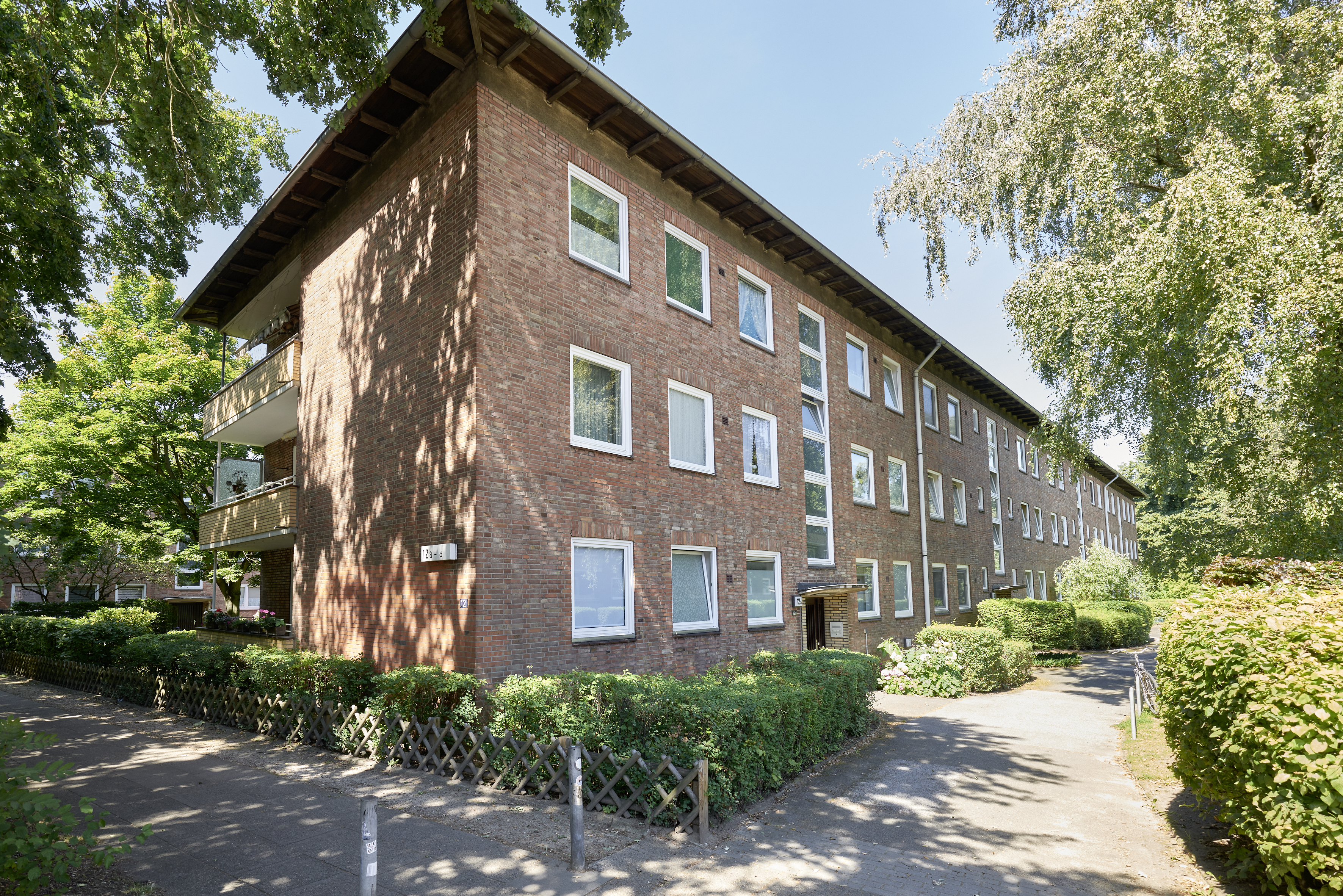 Dreigeschossiges Wohnhaus mit roter Backsteinfassade und Balkonen, umgeben von Bäumen, Hecken und begrünten Wegen.